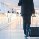 Businessman walking and wheeling a trolley suitcase at the lobby, talking on a mobile phone. Business travel concept.
