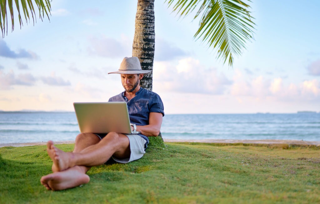 Individual sitting under a palm tree next to the ocean working on a laptop